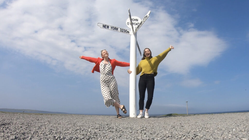 Two girls holding onto John O' Groats sign and smiling with arms up. NC500 .